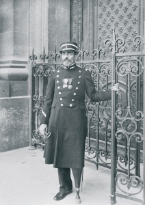 Georges Hector Dumont (1856-1930), gardien du tombeau de l’Empereur, devant le portail du Dôme des Invalides, vers 1910-1912. Il tient la clef du Dôme dans sa main gauche et porte une épée sur sa hanche gauche. © Paris, musée de l’Armée